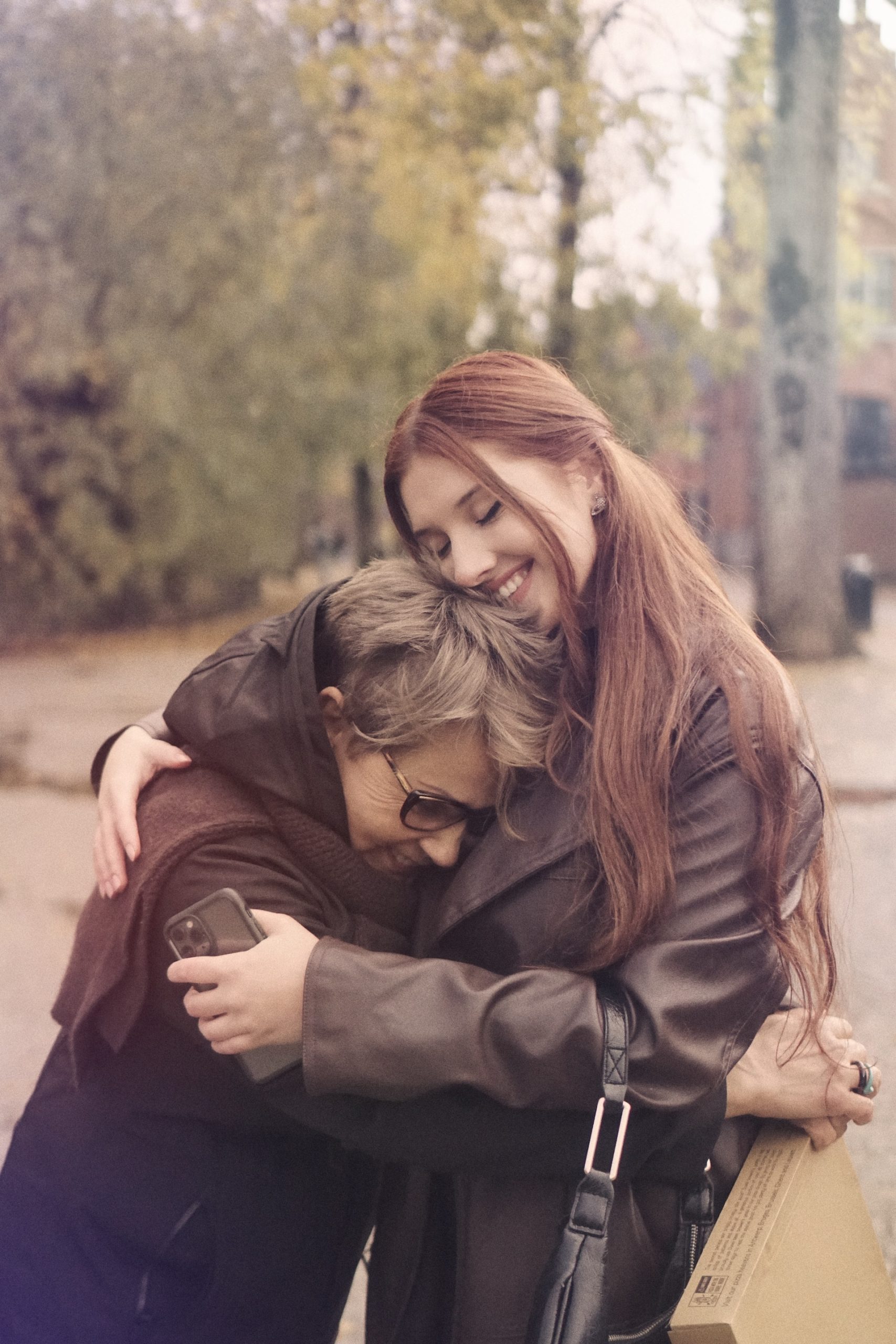 Young person hugging an older person and smiling. Foliage in the background.