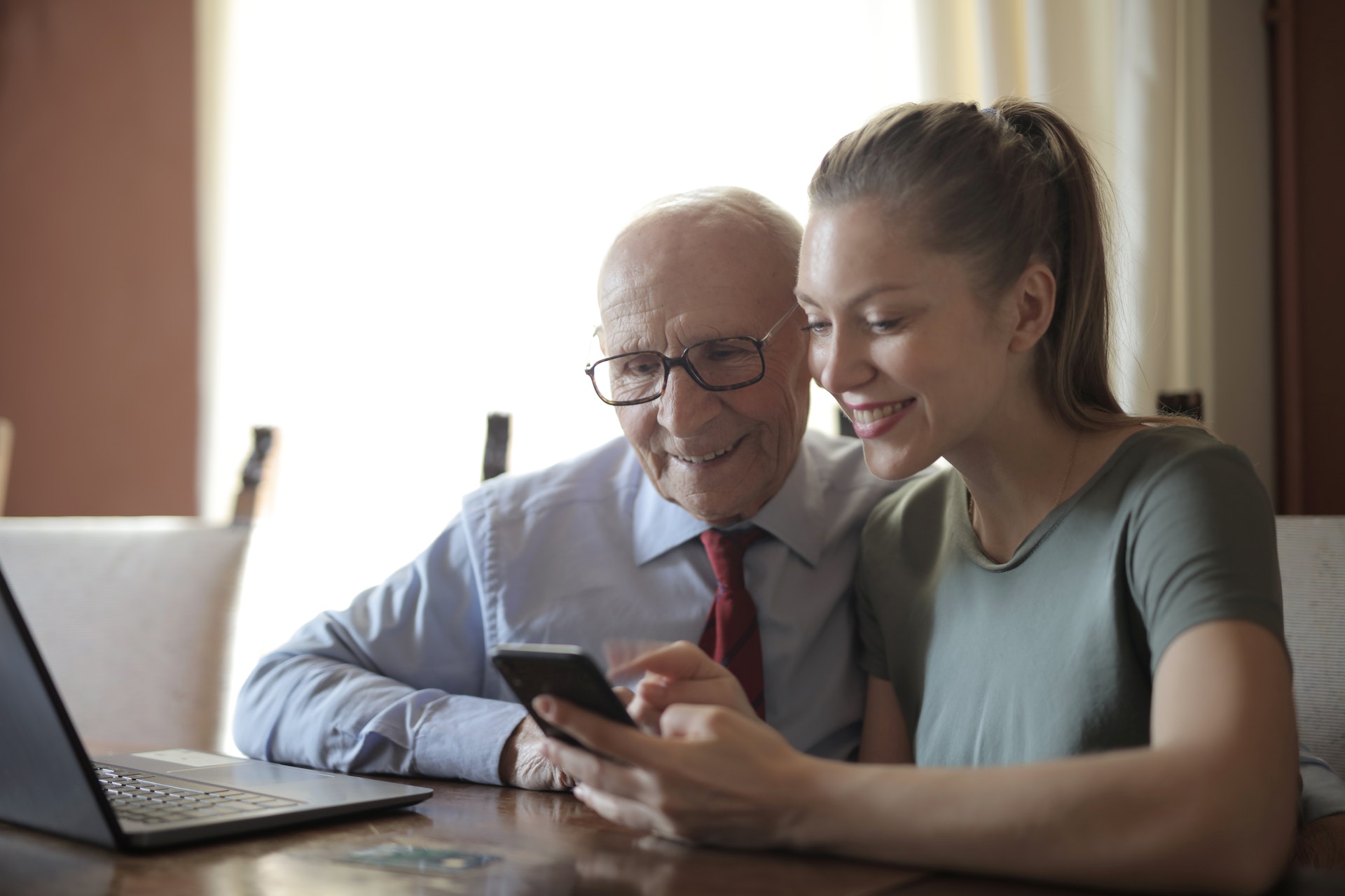 A young person showing her grandfather something on her phone as they sit in front of a computer and smile.