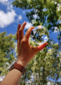 Image of a hand holding a clear item that shows the clouds, sky, and trees behind it.